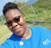 selfie of Lavern Ryan in a MapAction teeshirt outdoors with sea visible in the distance behind her